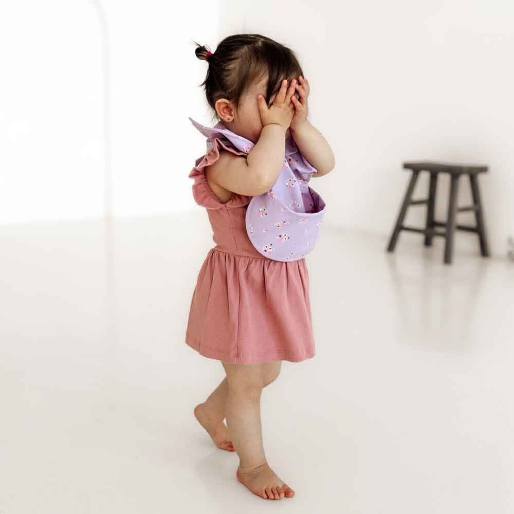 Baby girl in pink dress with lavender bib from Snuggle Hunny Lilac Bloom Frill Snuggle Bib
